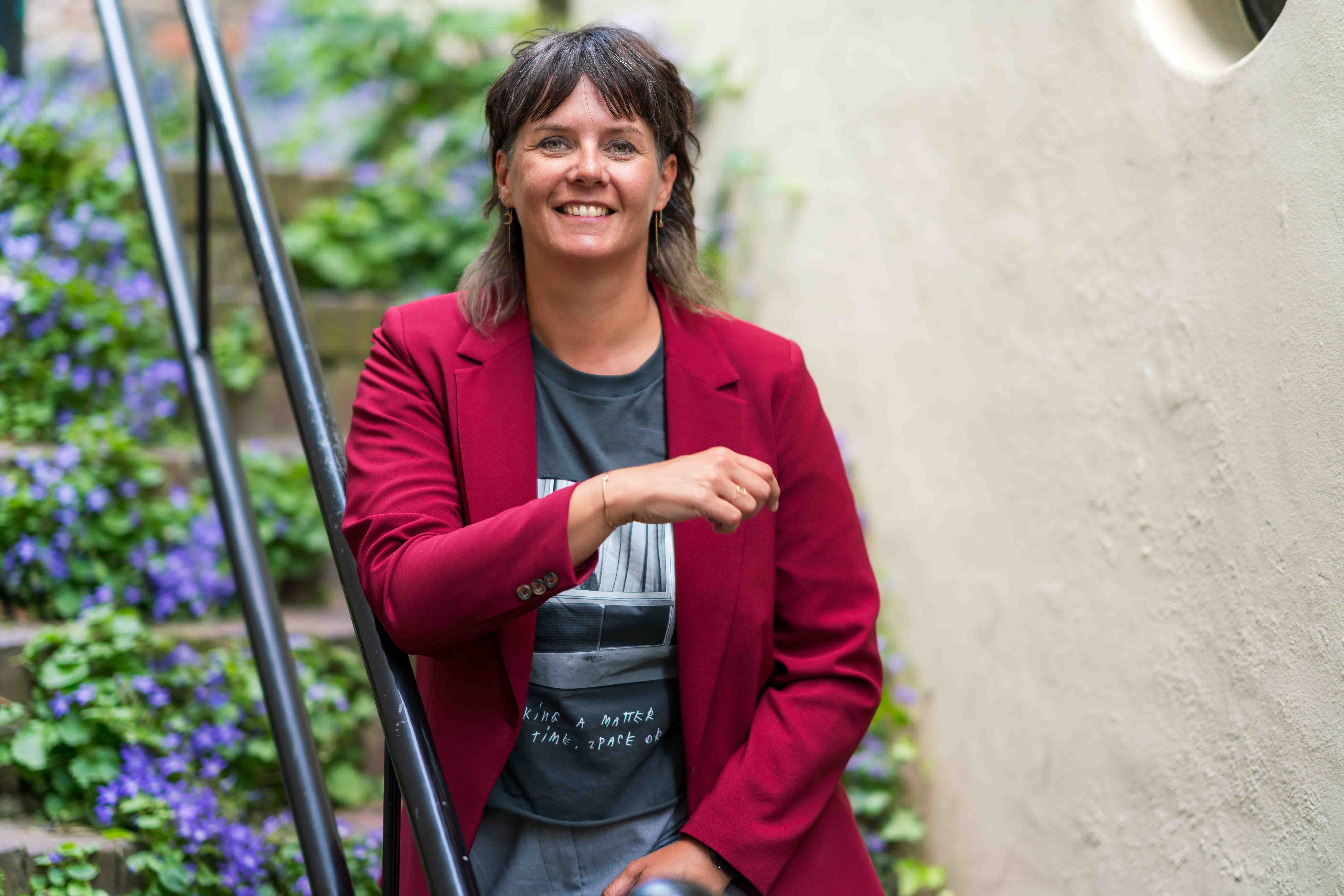 Saskia staand bij trapleuning met blauwe bloemen begroeiing op achtergrond